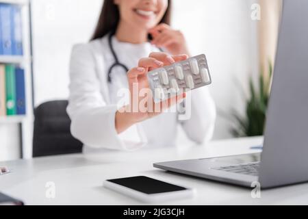 Cropped view of blurred doctor holding blister with pills during video call on laptop in hospital Stock Photo