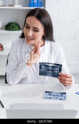 Positive doctor holding scan of teeth and pointing at cheek during video call on laptop in clinic Stock Photo