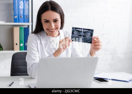 Positive doctor holding scan of teeth during video chat on laptop in hospital Stock Photo