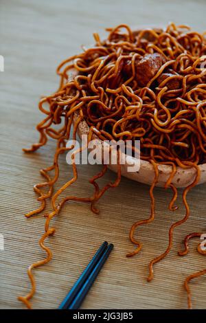 closeup of a brown ceramic plate with some chicken yakisoba noodles and a pair of black chopsticks placed on a table set with a beige tablecloth Stock Photo