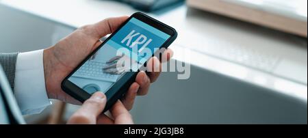 man at the office, wearing shirt and jacket, holding his smartphone, that reads the text KPI, for key performance indicator, in its screen, in a panor Stock Photo
