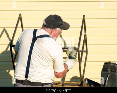 An senior gray-haired man processes a metal structure with a disc grinder. The master processes metal in the yard of a country house on a sunny summer day. Stock Photo