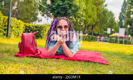 Young woman lying on a blanket records a voice message using a smartphone in the park. Happy girl lying on the grass and use voice recording app or di Stock Photo