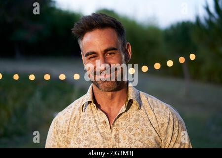 Portrait of happy mature man looking at camera outdoor. Senior people with beard feeling confident at sunset. Closeup face of smiling with backyard in Stock Photo