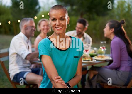 Portrait of a woman with shaved head looking at the camera. Group of friends dining outside on a terrace restaurant or backyard home. Friends having Stock Photo
