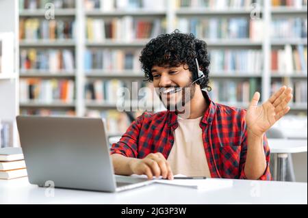 Cheerful positive Indian or Arabian male student or freelancer with headset, sitting at table in a library, using laptop, talking on video call, waving, listening to online webinar, lecture, smiling Stock Photo