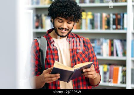 Focused clever positive indian or arabian university or college male student, standing in a library near bookshelf, reading interesting book, preparing for exam, doing homework, smiling Stock Photo