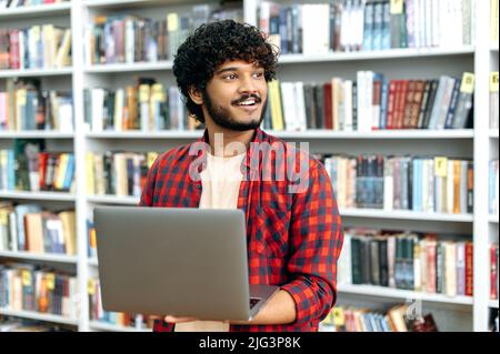 Positive indian or arabian curly haired guy, student, freelancer, in a casual shirt, holds an open laptop in his hands, stands in the library against the background of bookshelves, looks away, smiles Stock Photo