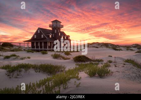 Oregon Inlet Lifesaving Station surrounded by sand dunes under a glowing purple, orange, and yellow sunset in the Outer Banks, North Carolina Stock Photo