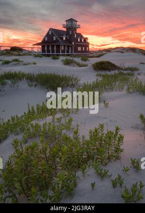 Oregon Inlet Lifesaving Station surrounded by sand dunes under a glowing purple, orange, and yellow sunset in the Outer Banks, North Carolina Stock Photo