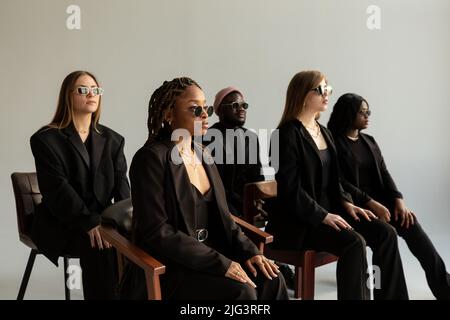 Five people of different nationalities sit on chairs and look in one direction. All dressed in black suits and glasses. Stock Photo