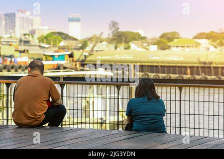 Back view of Asian couple relaxing and chilling at a riverside. Relaxed lifestyle for lover enjoying freedom in summer holidays. Happy people outdoors Stock Photo