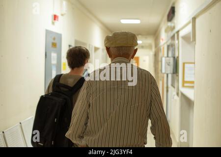 Pensioner in hospital. Grandpa in hallway of medical facility. Elderly person is looking for doctor's office. Grandson takes grandfather for examinati Stock Photo