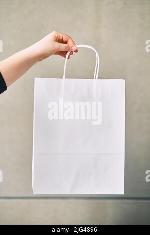 Young woman holding white paper disposable bag in hand on gray background  Stock Photo