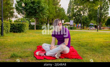 A handsome guy with dreadlocks in casual clothes, sitting on a plaid in a park on a green lawn and looking thoughtfully into the distance. A handsome Stock Photo