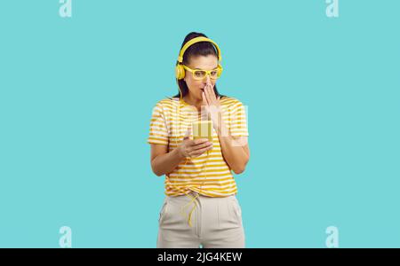 Young beautiful woman with headphones watching something incredible on the phone covers mouth in shock, looks shy, standing on blue background. Stock Photo