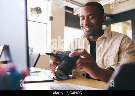 Young Black Professional Photographer Sitting At Desk Working On Computer Holding Camera Editing Pictures Stock Photo