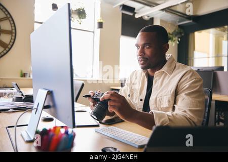 Young Black Professional Photographer Sitting At Desk Working On Computer Holding Camera Editing Pictures Stock Photo