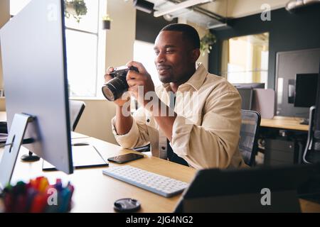 Young Black Professional Photographer Sitting At Desk Working On Computer Holding Camera Editing Pictures Stock Photo