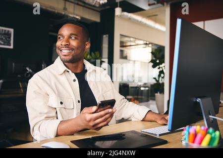 Young Black Male Smiling Advertising Marketing Or Design Creative In Modern Office Sitting At Desk Using Mobile Phone Stock Photo