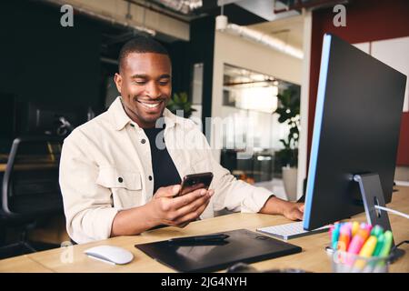 Young Black Male Advertising Marketing Or Design Creative In Modern Office Sitting At Desk Using Mobile Phone Stock Photo