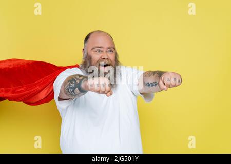 bearded overweight man in superhero costume shouting with outstretched hands   isolated on yellow Stock Photo