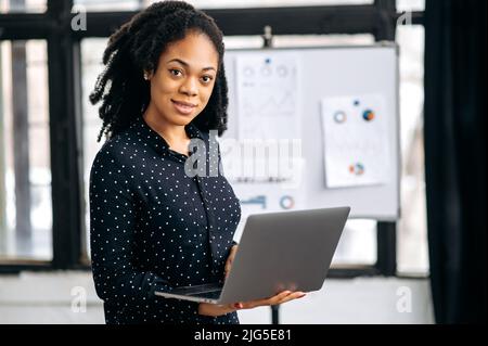 Portrait of a beautiful positive african american young woman, in formal shirt, an office worker, top manager, holds an open laptop in her hands, stands in a modern office, looks at the camera, smiles Stock Photo