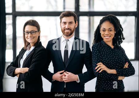 Portrait of successful group of multiracial business people in a modern office, looking at camera, smiling. Happy proud office employees, a man and two women, standing in meeting room of their office Stock Photo