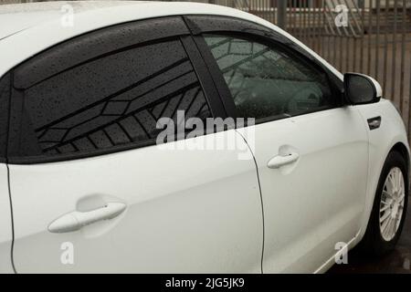 White car in parking lot in rain. Transport in city. Black glass in car. Tinted glass. Stock Photo