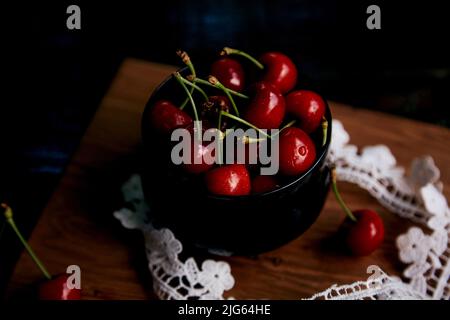 Fresh cherry in glass bowl on the sacking on black background in a low key with lace decoration. Rustic organic home-grown berries. Cozy countryside dinner. Stock Photo