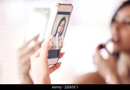I love the way this camera makes me look. Shot of a beautiful young woman using her phone as a mirror while applying lipstick at home. Stock Photo