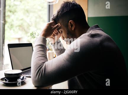 .. Stressed young mixed race man sitting at a table in a cafe drinking coffee and using a laptop alone. Unhappy hispanic businessman sitting in a Stock Photo
