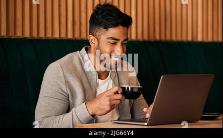 .. Happy young mixed race man sitting at a table in a cafe drinking coffee and using a laptop alone. One hispanic businessman sitting in a restaurant Stock Photo