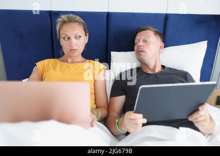 Couple working using gadgets in bed, man prying about woman Stock Photo