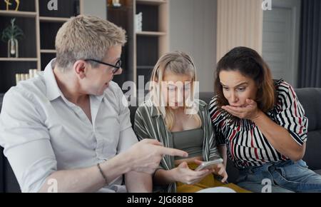 Caucasian parents sitting on sofa and excited daughter using smart phone, they're happy and wondering in something on screen with strange face in livi Stock Photo