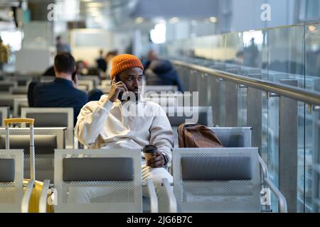 African traveler man drinking coffee waiting for flying at airport terminal, talking on cellphone. Stock Photo