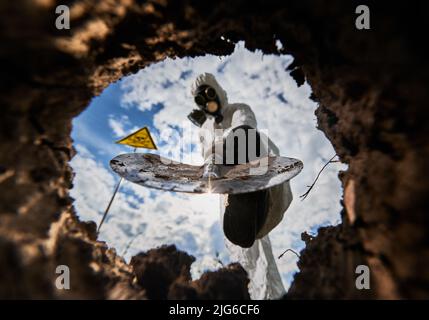 Close up view from inside pit of shovel digging by person in protective coverall and gas mask, on backdrop of warning sign with skull with crossed bones. Stock Photo