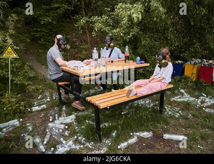 Man, woman and child in gas masks sitting at the table in forest with trash plastic bottles and poison toxic sign. Family resting in the woods with garbage and skull-and-crossbones warning symbol. Stock Photo