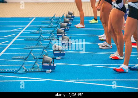 Track and field, running and sprinting. Legs of professional athletes before the start of a sprint race. Female athletes stand on the blue track befor Stock Photo