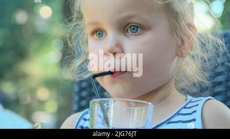 Cute little girl drinks juice through straw. Close-up portrait of blonde girl drinks juice from glass through cocktail straw sitting in street cafe Stock Photo