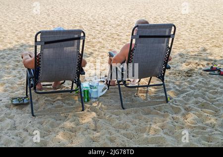Two men sitting sunbathe in sun loungers on river beach under the shade of tree Stock Photo