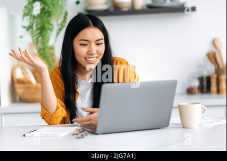 Online video communication. Friendly pretty asian brunette young woman, manager, freelancer, sit in the kitchen, talking on video call by laptop with the client or employees, discussing ideas, smiling Stock Photo