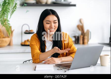 Virtual meeting, briefing. Happy chinese brunette young woman, freelancer, company employee, sit in the kitchen, talking on video call by laptop with the client or employees, discussing ideas, smiling Stock Photo