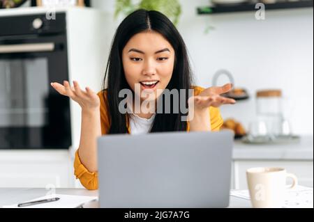 Excited positive Chinese young woman, student, freelancer, sits at home in the interior kitchen, emotionally talking on video conference with friends, employees, customers, gesturing with hands, smile Stock Photo