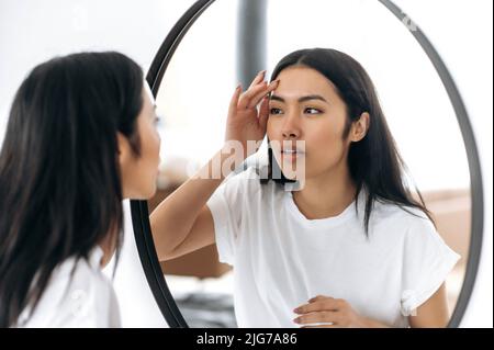 Satisfied lovely chinese girl with long brunette hair in white t-shirt, stands in front of a mirror, examines her face, happy at the healthy state of her facial skin, smile, getting ready to do makeup