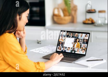Remote lesson. Positive smart Asian female student, sitting at the table, listening to an online lecture, on the screen of a laptop, a teacher telling information, and a group of multiracial people Stock Photo