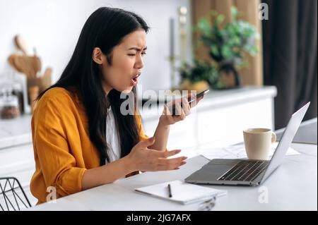Annoyed asian young woman, freelancer, student, ceo, sits at home in the kitchen, work remotely, holding smart phone in hand speak on speakerphone, record audio message, emotional face expression Stock Photo