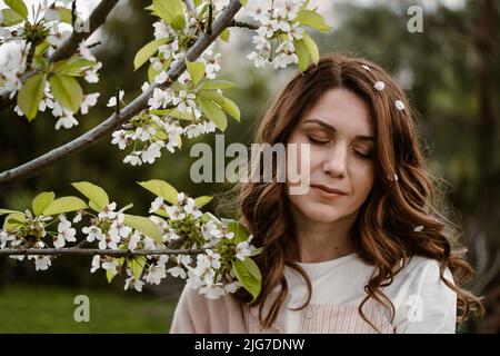 Beautiful adult woman closed eyes, enjoying flowers and smelling blossom tree in the spring garden. Close-up portrait with natural makeup Stock Photo