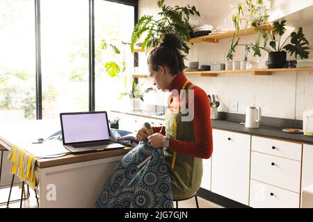 Image of caucasian non-binary trans woman with laptop working in sewing workshop Stock Photo
