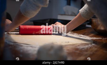 Low angle view of a woman using red rolling pin to roll homemade vegan pastry dough for strudel or pie on a domestic dining table. Stock Photo